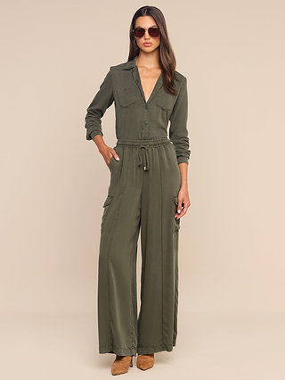 A woman with brown hair, sunglasses, and brown heels models the Chandler Cargo Pant in olive green. She is styled in a long-sleeve jumpsuit featuring a drawstring waist, chest pockets, and wide-leg silhouette, posing against a plain beige background.