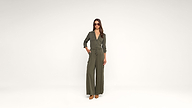 A woman wearing sunglasses and an olive green jumpsuit with long sleeves and wide legs stands confidently against a plain white background, with one hand in her pocket and brown shoes visible.