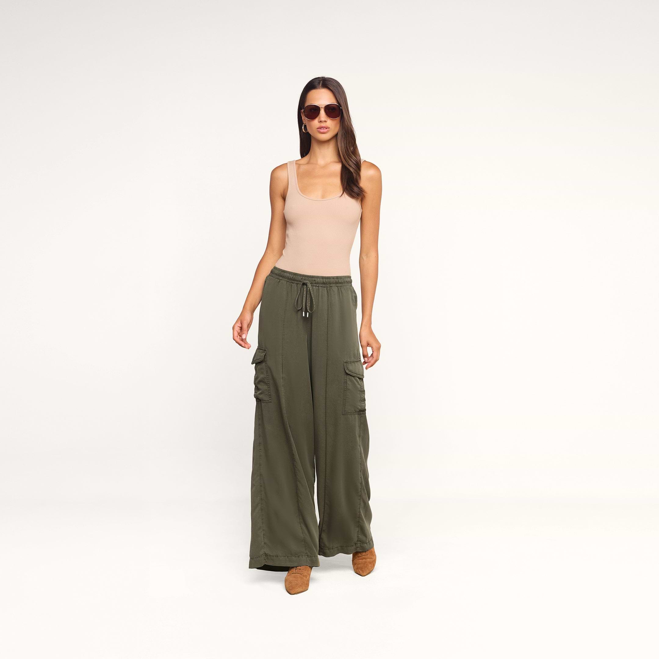 A woman stands against a plain white background wearing sunglasses, a beige tank top, brown shoes, and the Chandler Cargo Pant in loose olive green wide-leg style.