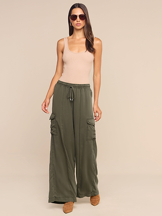 A woman poses against a neutral background wearing sunglasses, a beige tank top, brown shoes, and the Chandler Cargo Pant—a loose-fitting olive green wide-leg style with pockets.