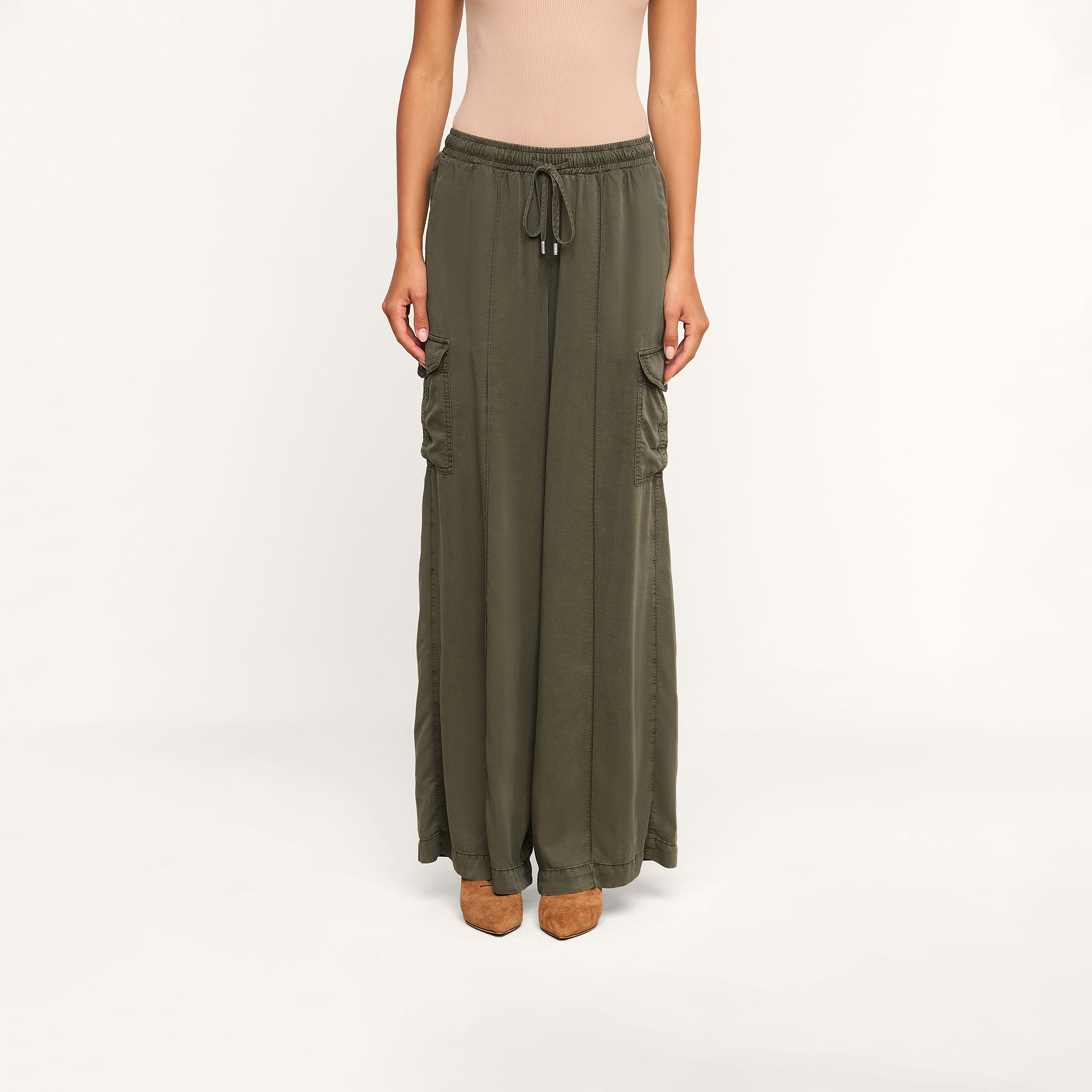 Wearing the Chandler Cargo Pant—a pair of olive green, wide-leg cargo pants with side pockets and a drawstring waist—alongside a beige sleeveless top and brown heeled shoes, a person stands against a plain white background.