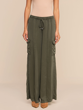 A person wearing the Chandler Cargo Pant—olive green, wide-leg cargo pants featuring ankle-length hems, a drawstring waist, and side pockets—paired with a light beige sleeveless top and brown shoes, stands against a plain background.