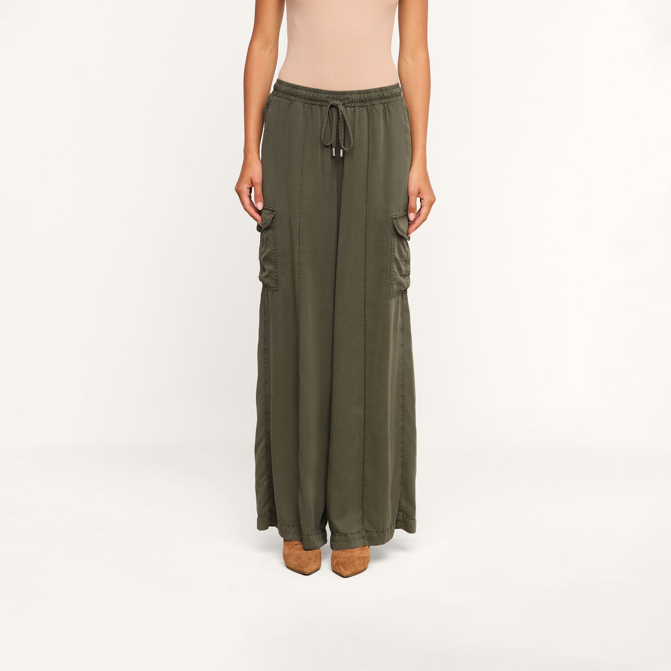 Wearing the Chandler Cargo Pant—a pair of olive green, wide-leg cargo pants with side pockets and a drawstring waist—alongside a beige sleeveless top and brown heeled shoes, a person stands against a plain white background.