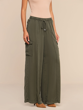 Wearing the Chandler Cargo Pant in olive green, featuring wide legs, large side pockets, and a drawstring waist, paired with a sleeveless beige top and brown pointed-toe heels, the person stands against a plain beige background.