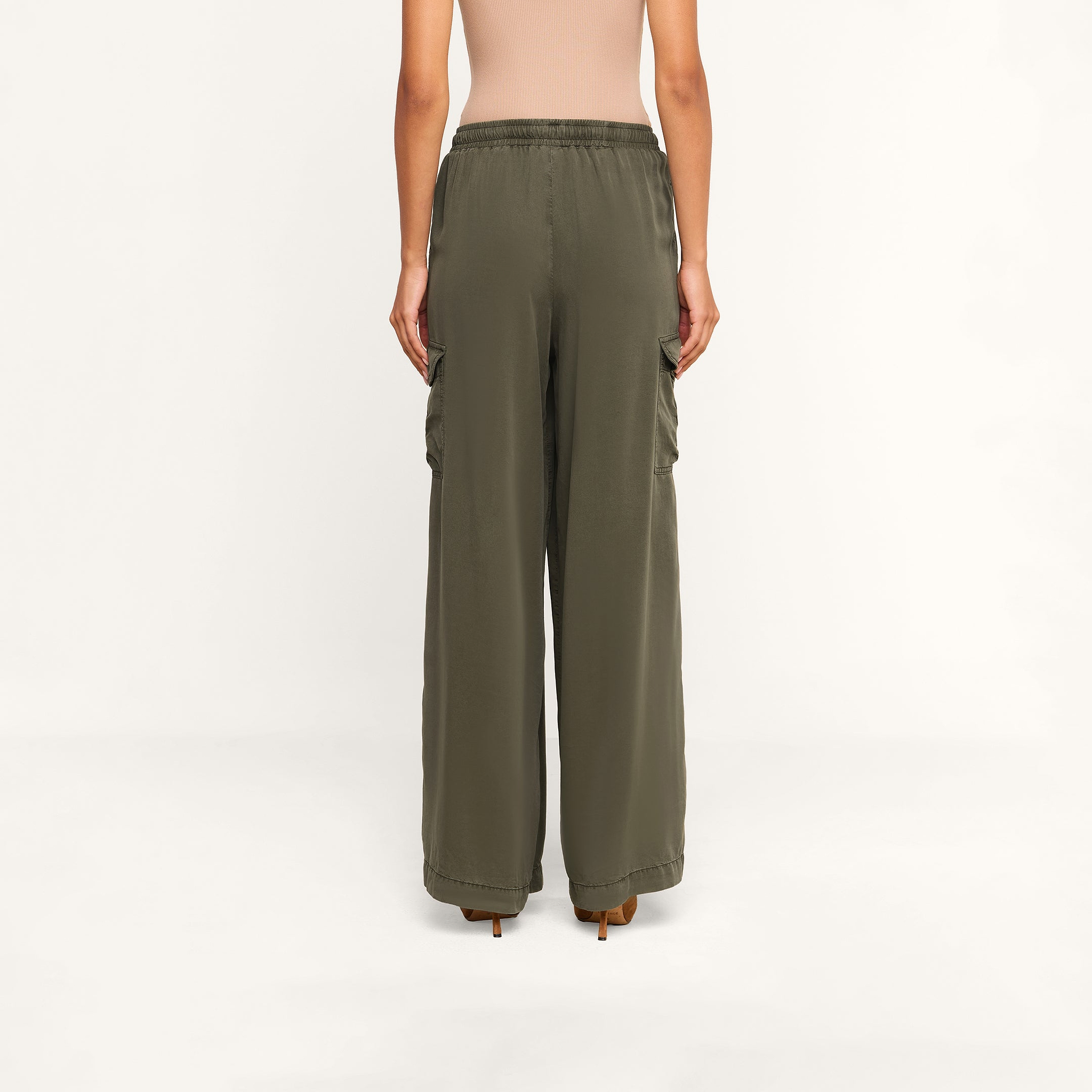 A woman stands facing away, wearing the Chandler Cargo Pant in olive green, styled with a beige top. These wide-leg cargo pants feature an elastic waistband and side pockets as she poses against a plain white background.