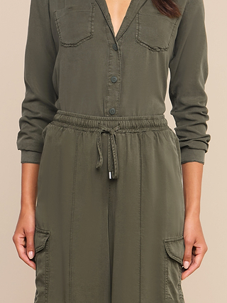 A person wearing the Chandler Cargo Pant in olive green, featuring a button-up design with long sleeves, chest pockets, a drawstring waist, and wide-leg styling. Only the body is visible from neck to mid-thigh.
