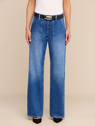 A person wearing the Christel Wide-Leg Jean, a high-rise, wide-leg style crafted from all-cotton denim with front patch pockets, styled with a black belt and black pointed-toe shoes. The image displays only the torso and legs against a plain beige background.