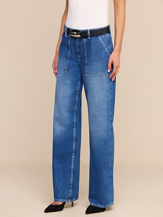 Someone is wearing the Christel Wide-Leg Jean, high-waisted and wide-leg blue jeans made from 100 cotton denim with front patch pockets. The outfit includes a black belt, black pointed-toe heels, and a sleeveless white top. The background is plain beige.