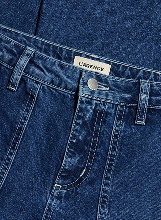 Close-up of blue denim Christel Wide-Leg Jean featuring white stitching, a front button, part of a pocket, and a white label with the brand name L’AGENCE sewn onto the inside waistband.