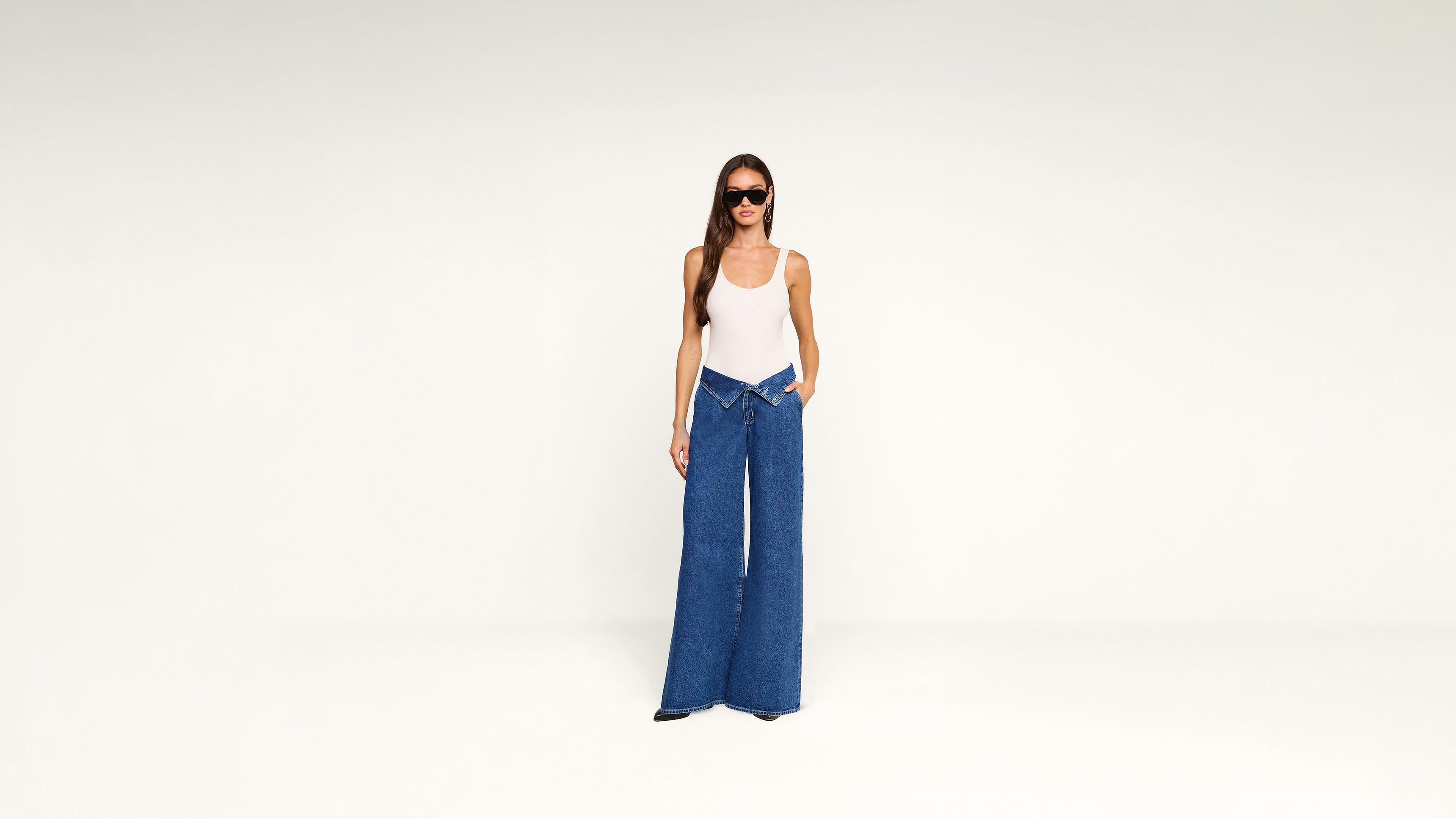 A woman wearing a white tank top, dark sunglasses, and blue wide-leg jeans with a folded waistband.