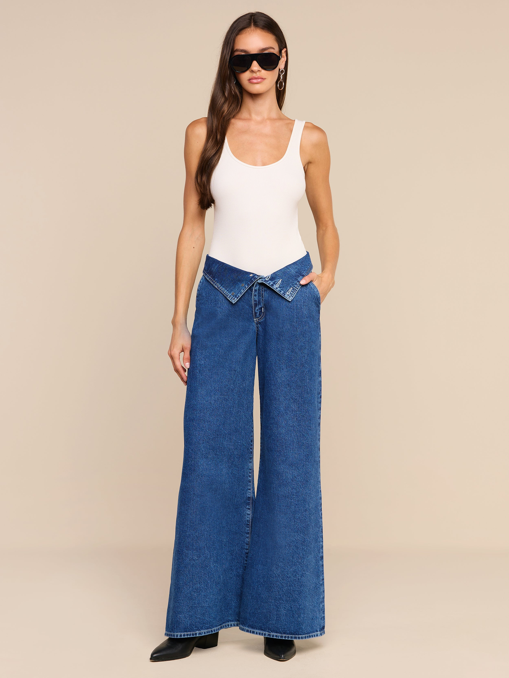 A woman wearing black sunglasses, a white tank top, and the Sonja Corset Jean—an ultra high-rise wide-leg blue jean with a corset waistband—stands against a plain white background.