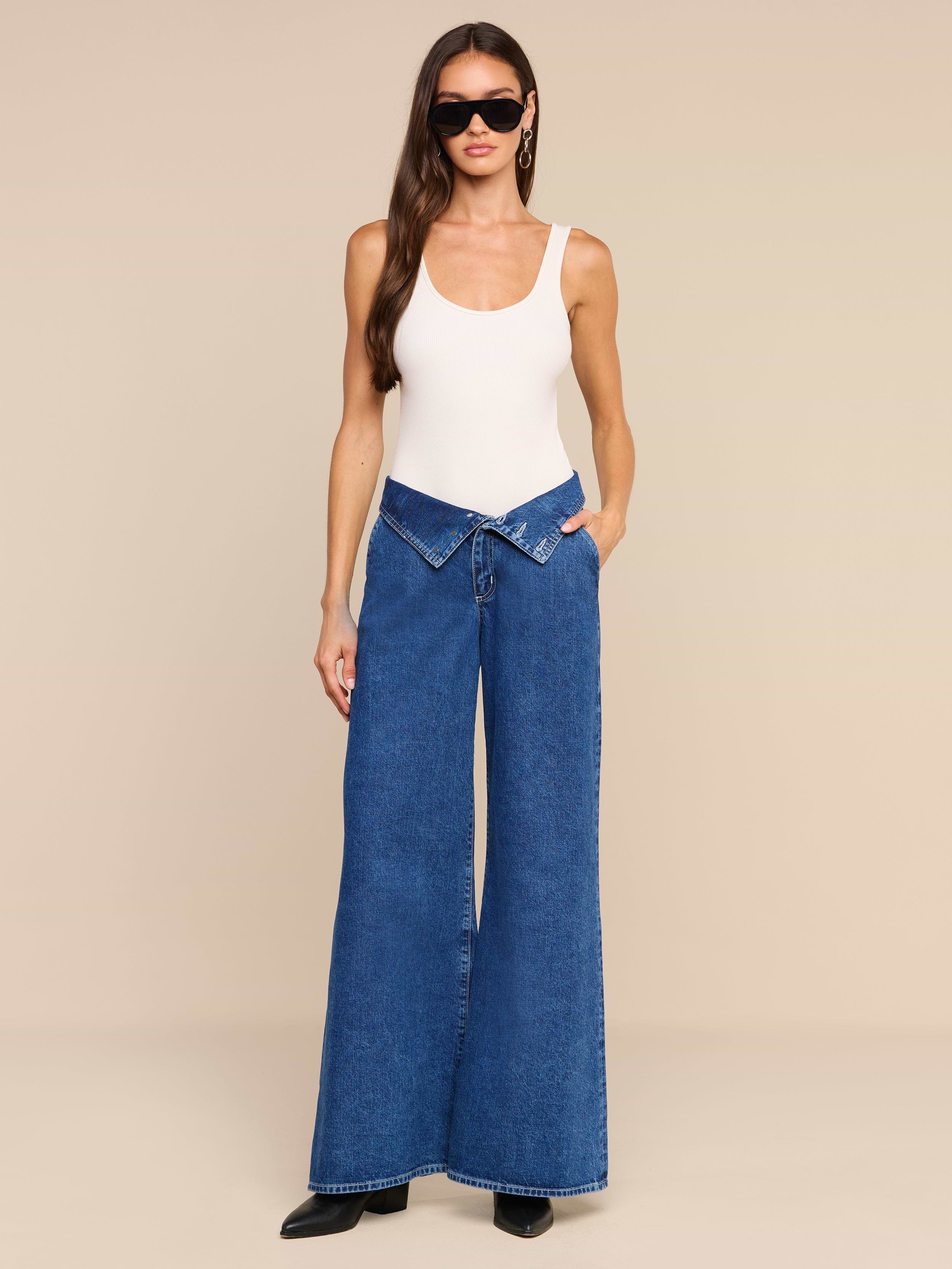 A woman wearing black sunglasses, a white tank top, and the Sonja Corset Jean—an ultra high-rise wide-leg blue jean with a corset waistband—stands against a plain white background.