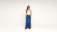 A woman in a white tank top, wide-leg blue jeans with a V-shaped waistband, black shoes, and sunglasses stands against a plain white background.