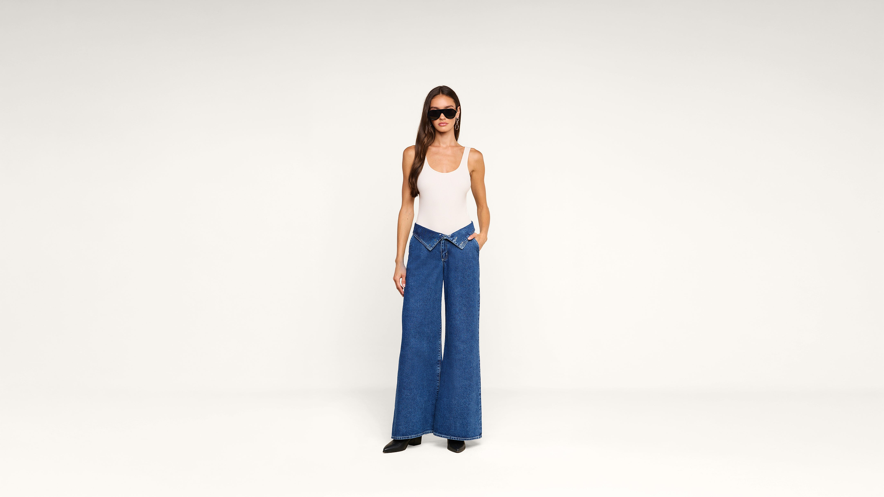 A woman in a white tank top, wide-leg blue jeans with a V-shaped waistband, black shoes, and sunglasses stands against a plain white background.