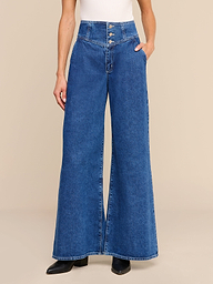 A person is wearing the Sonja Corset Jean—an ultra high-rise, wide-leg jean with a corset waistband—paired with a white sleeveless top and black shoes, standing against a plain white background.