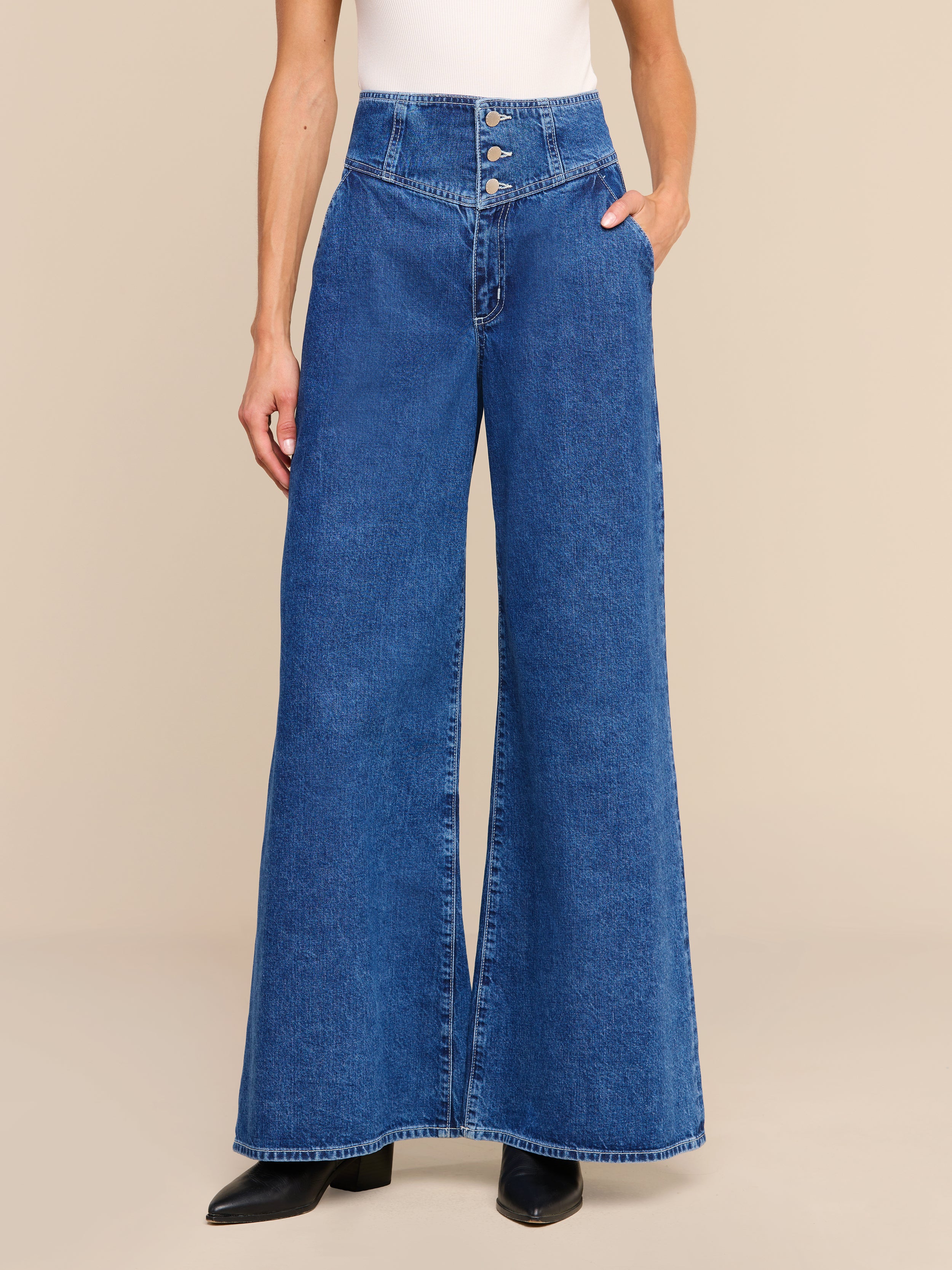 A person is wearing the Sonja Corset Jean—an ultra high-rise, wide-leg jean with a corset waistband—paired with a white sleeveless top and black shoes, standing against a plain white background.