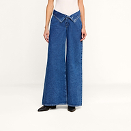 A person wearing the Sonja Corset Jean—ultra high-rise wide-leg jeans featuring a V-shaped crossover waistband—with a white top and black boots stands against a plain white background.
