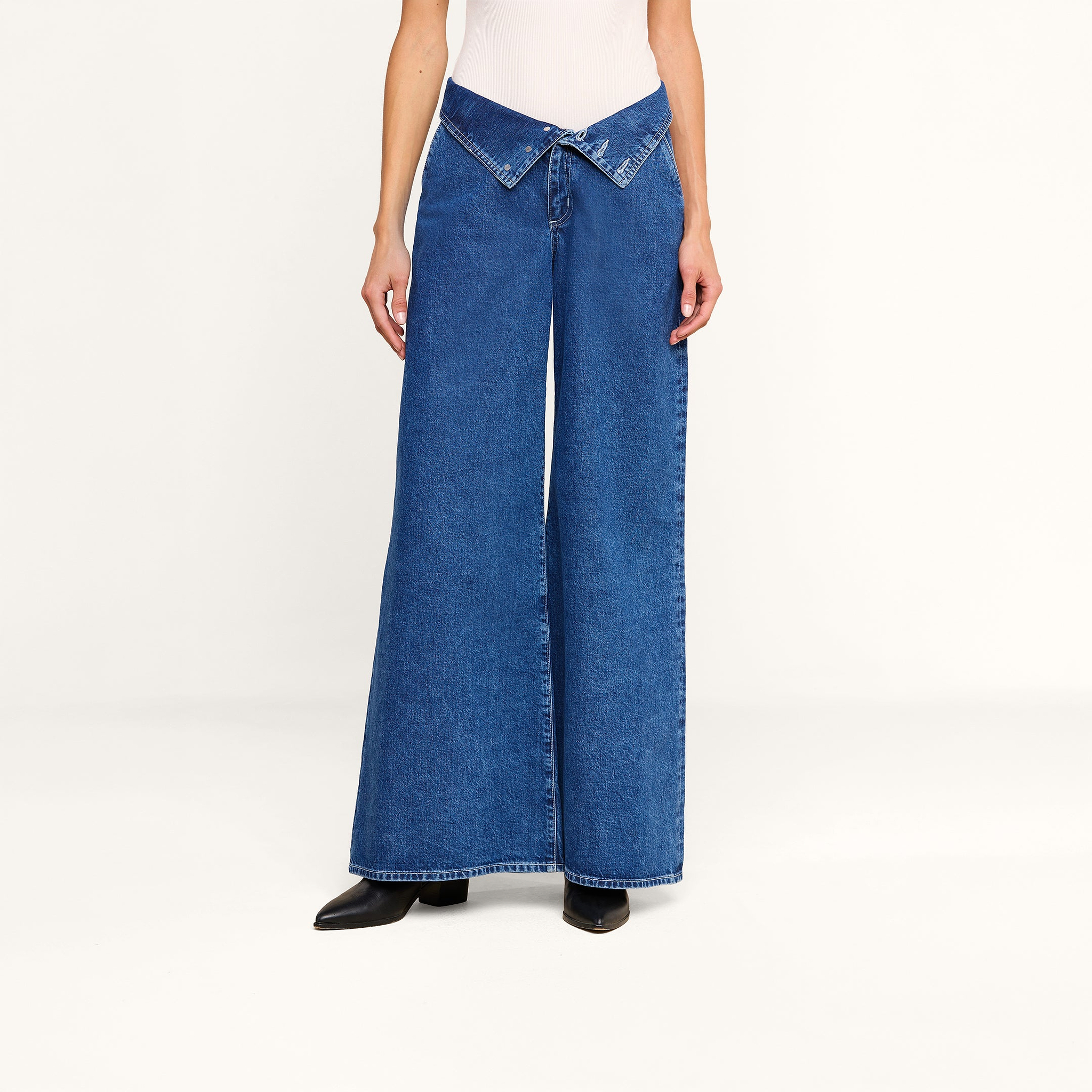 A person wearing the Sonja Corset Jean—ultra high-rise wide-leg jeans featuring a V-shaped crossover waistband—with a white top and black boots stands against a plain white background.