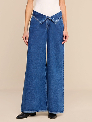Wearing the Sonja Corset Jean—an ultra high rise, wide-leg jean featuring a corset-inspired waistband—a person stands against a plain beige background. The look is styled with a white top and black pointed-toe shoes.