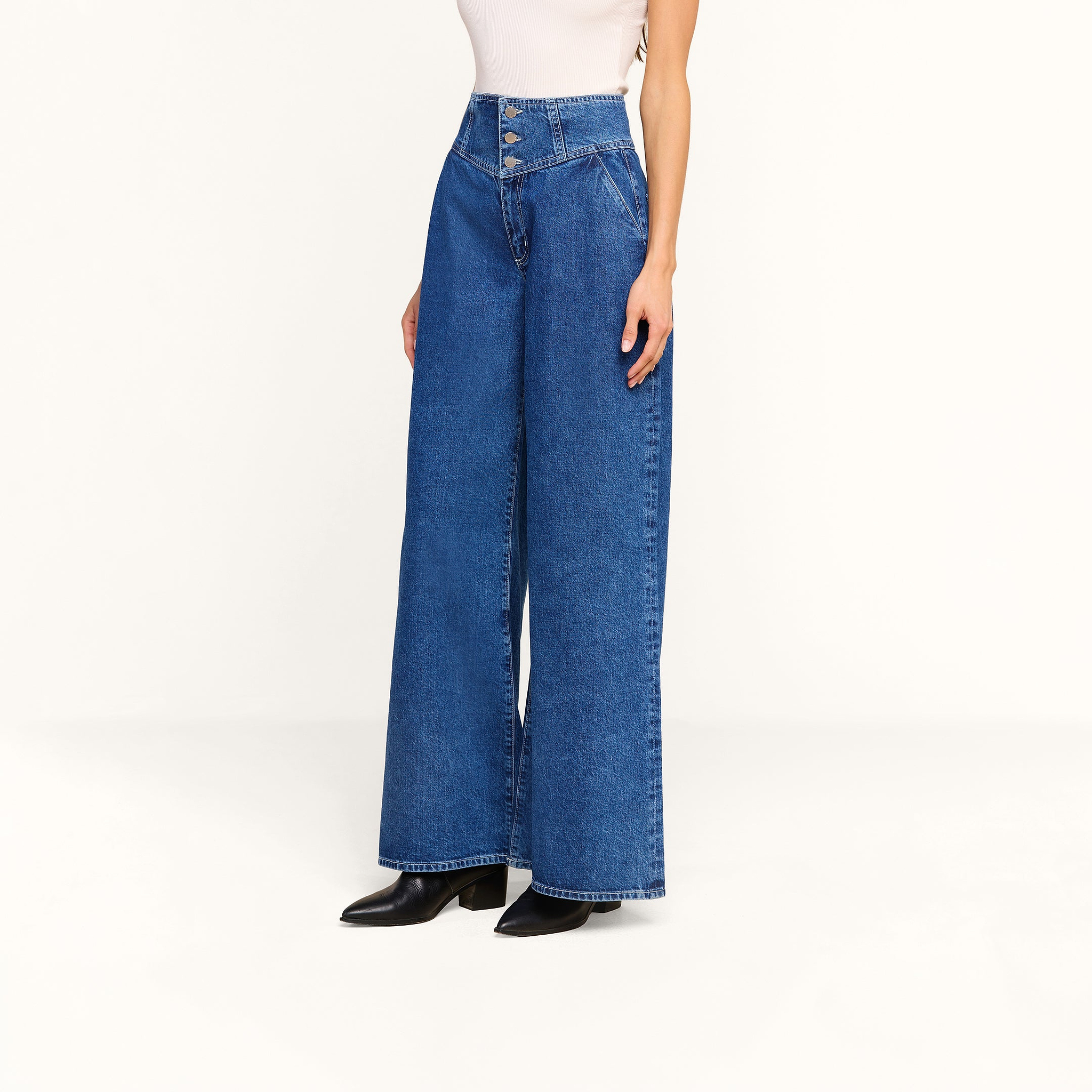 A person wearing the Sonja Corset Jean—ultra high-rise, wide-leg blue jeans—with a fitted white top and black pointed-toe shoes stands against a plain white background.