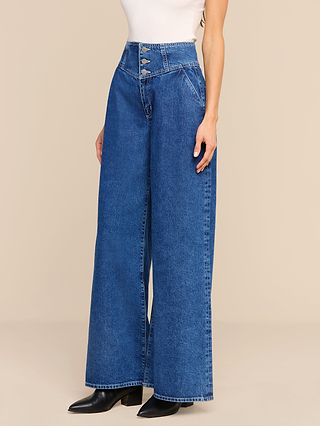 Wearing the Sonja Corset Jean—an ultra high rise, wide-leg blue jean featuring a corset-inspired waistband and multiple front buttons—a woman stands against a beige background, paired with a white sleeveless top and black pointed-toe boots.