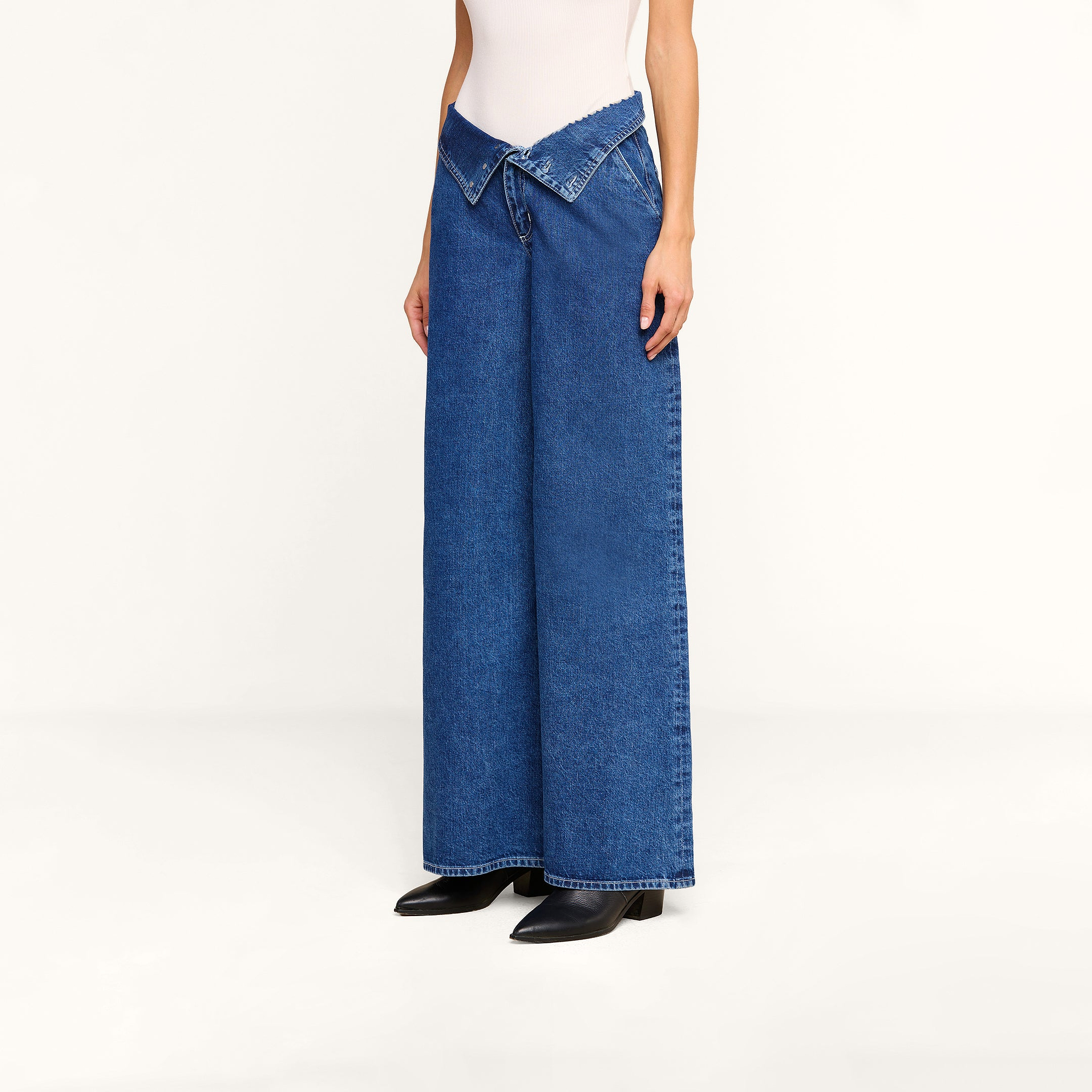 Wearing the Sonja Corset Jean, featuring an ultra high-rise wide-leg silhouette and a distinctive corset waistband in blue, styled with a white top and black pointed-toe shoes against a plain white background.