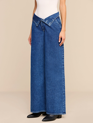 Person wearing the Sonja Corset Jean—ultra high rise, wide-leg blue jeans with a corset-inspired waistband and a distinctive V-shaped, folded-over detail—styled with a white sleeveless top and black boots, set against a beige background.