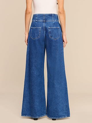A person is shown from the back against a plain beige background wearing the Sonja Corset Jean—an ultra high-rise, wide-leg blue jean with a subtle corset-inspired waistband—and a white sleeveless top.