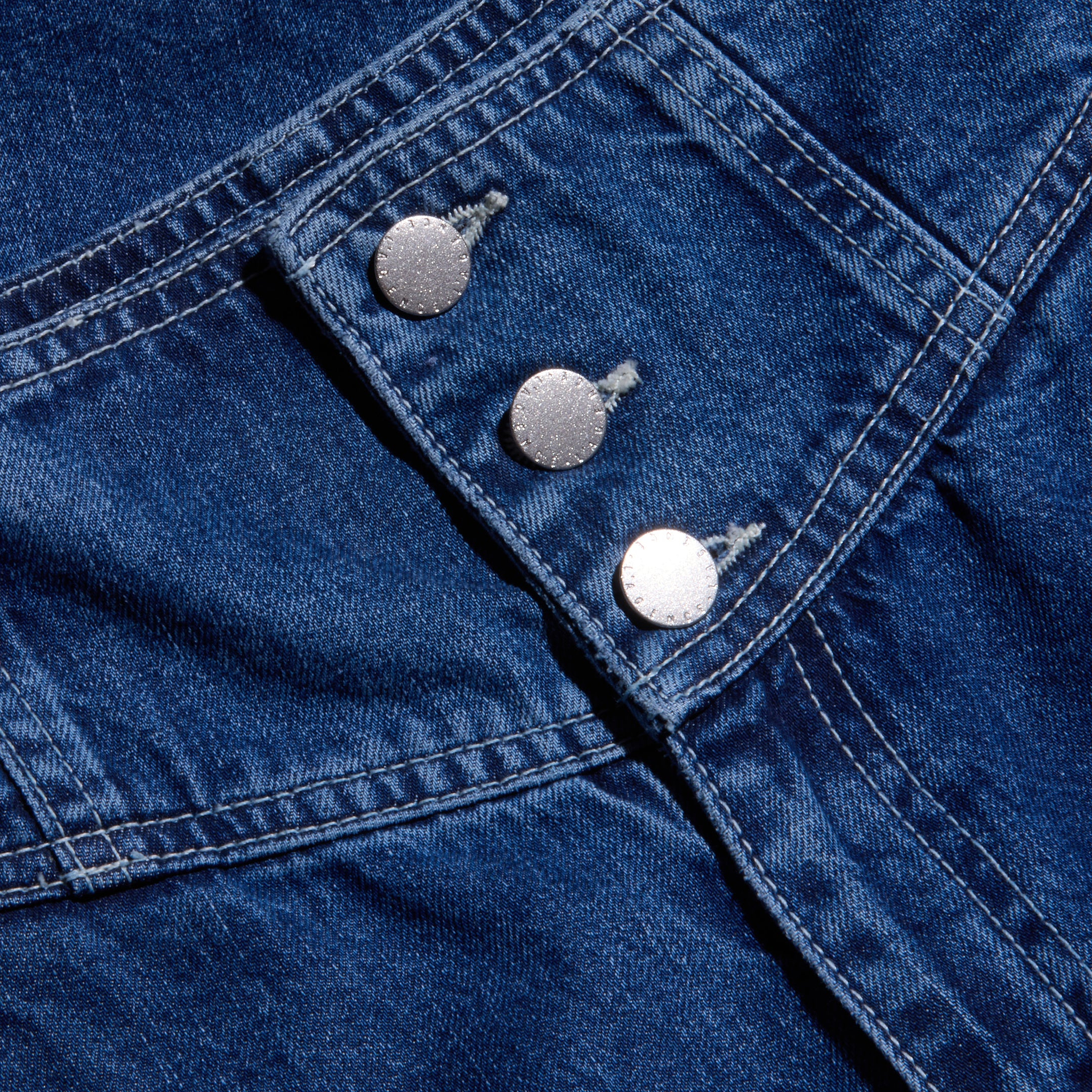 Close-up of the Sonja Corset Jean in blue denim, featuring three silver buttons, visible stitching, and diagonal seam lines on an ultra high-rise corset waistband. The texture of the material and the shine on the buttons are clearly shown.