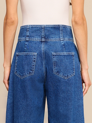 A person wearing the Sonja Corset Jean in ultra high-rise blue denim, featuring a corset-inspired waistband, paired with a white ribbed tank top, shown from the back with arms relaxed by their sides.