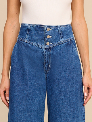 Wearing the Sonja Corset Jean—an ultra high rise blue denim jean with a corset-inspired waistband, three silver buttons, and front pockets—this look is paired with a white ribbed sleeveless top for a modern, wide-leg silhouette.