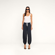 A woman wearing sunglasses, a white sleeveless top, and the Mariko Jogger black lightweight nylon pants stands facing forward against a plain white background. She completes her look with black high-heeled sandals.