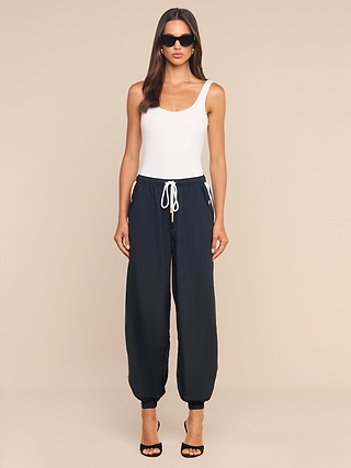 A woman wearing a white tank top, the Mariko Jogger in black—crafted from lightweight nylon with a water repellent finish—black high-heeled sandals, and sunglasses stands against a plain beige background.