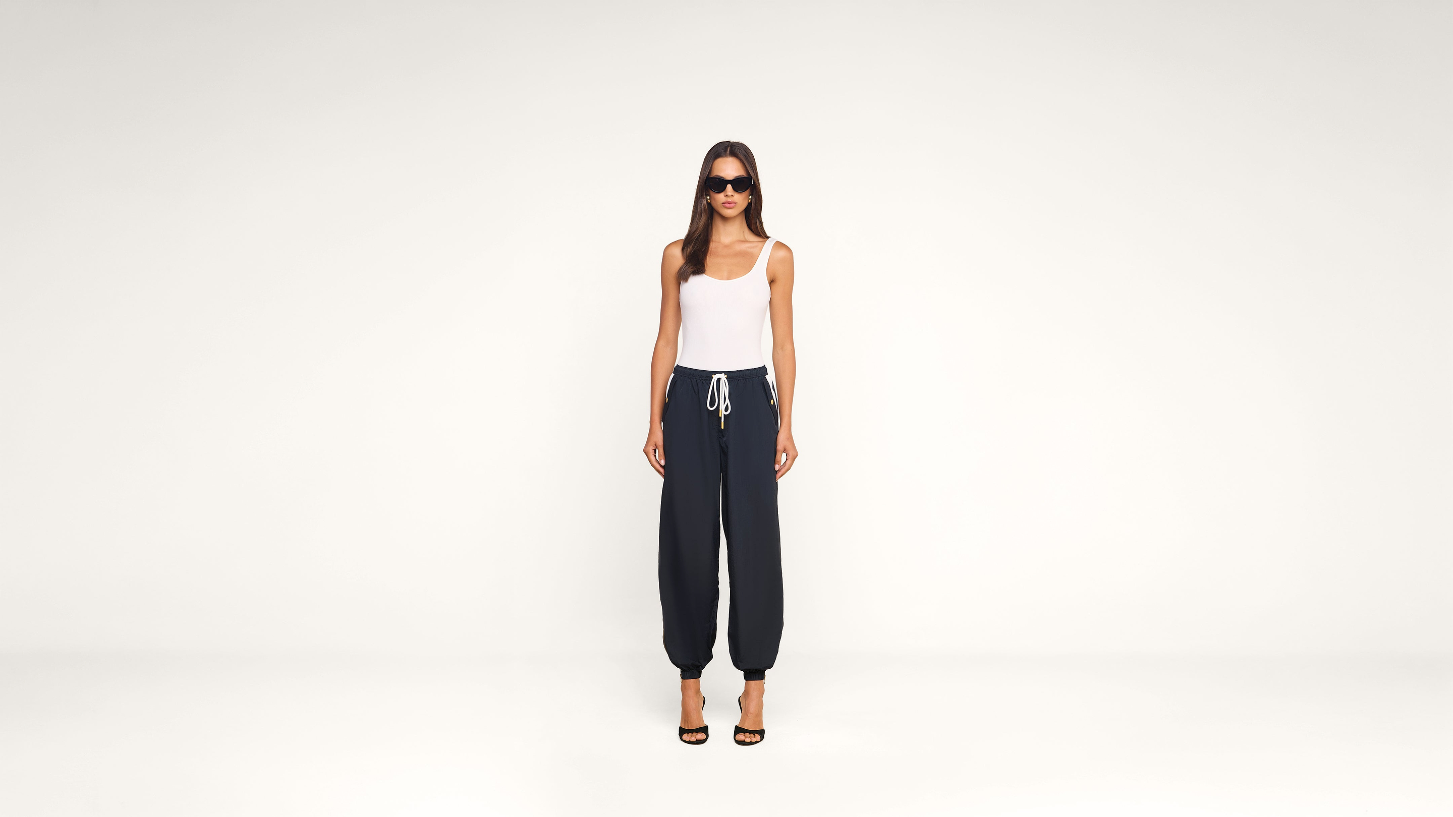 A woman stands against a plain white background, wearing sunglasses, a white tank top, dark drawstring pants, and black high-heeled sandals.