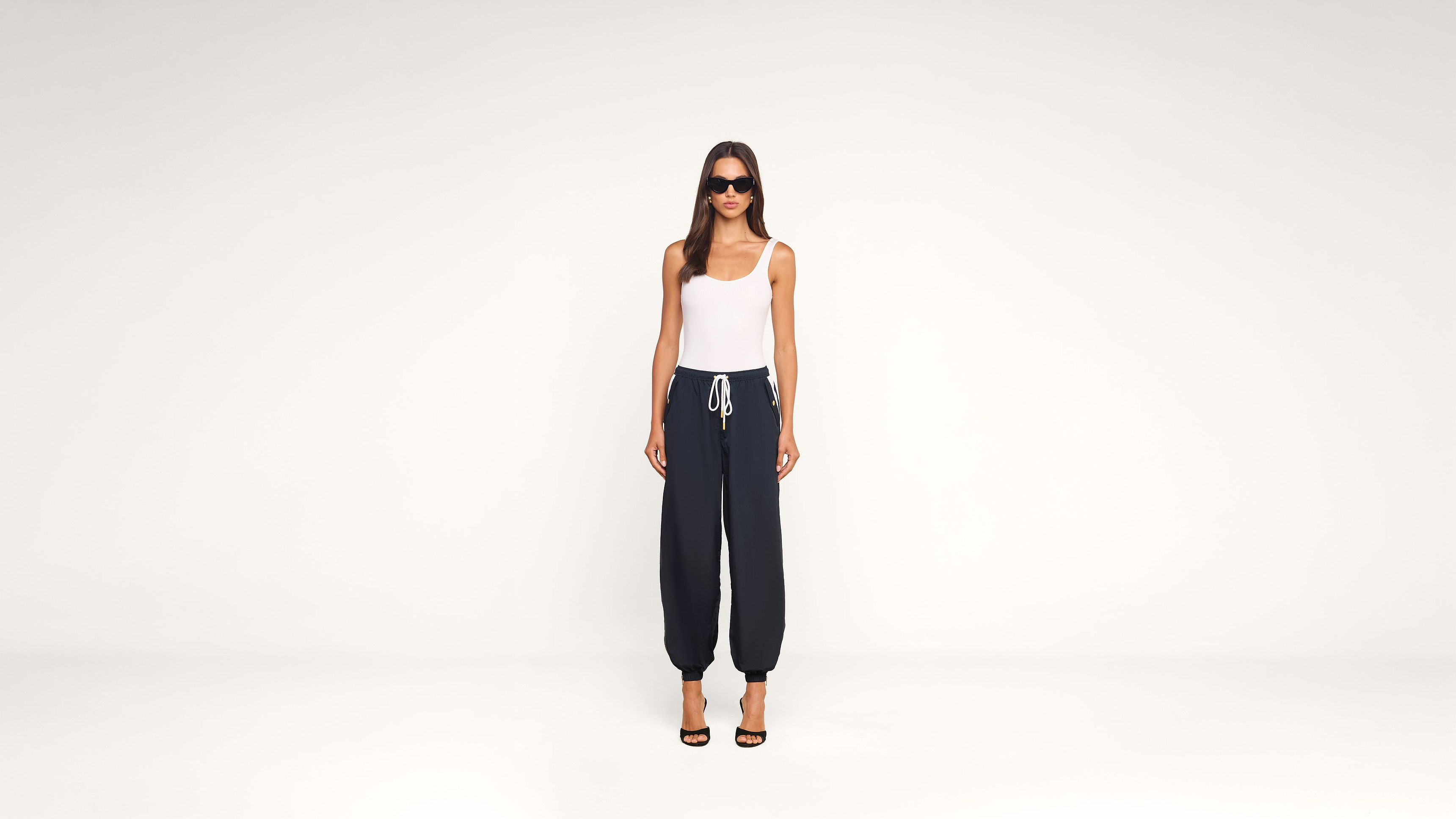 A woman stands against a plain white background, wearing sunglasses, a white tank top, dark drawstring pants, and black high-heeled sandals.