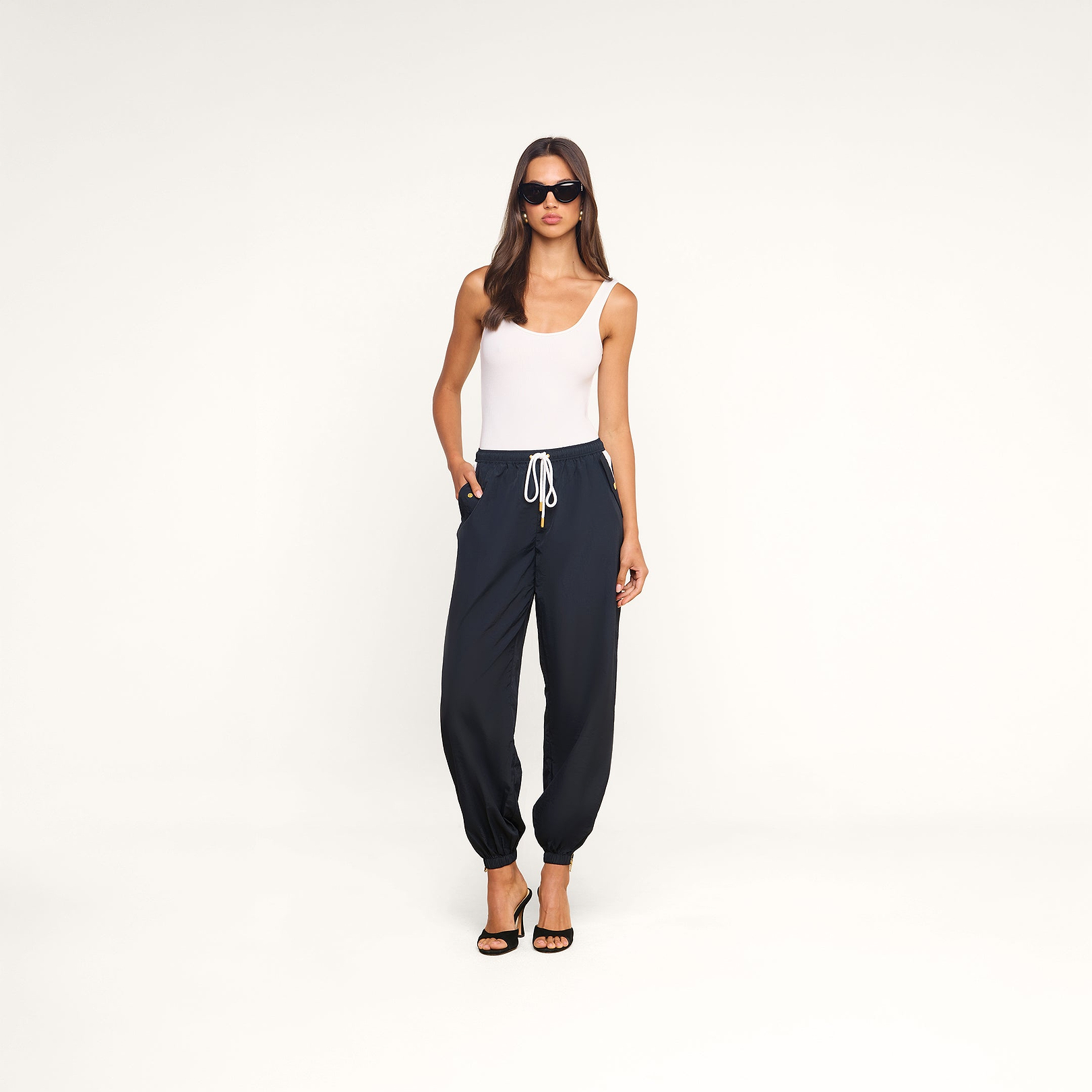 A woman in sunglasses wears a white tank top, the navy blue Mariko Jogger made from lightweight, water-repellent nylon, and black heeled sandals while standing against a plain white background.