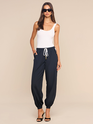 A woman stands against a plain beige background, hands in her pockets, wearing sunglasses, a white sleeveless top, black heeled sandals, and the Mariko Jogger—navy jogger pants crafted from lightweight nylon with a water-repellent finish and a drawstring.