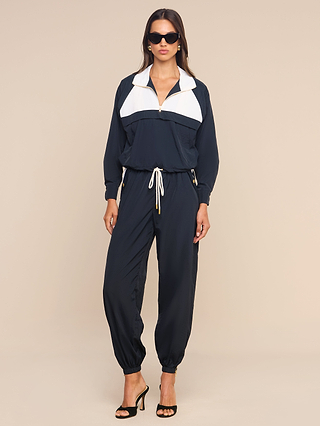 A woman stands against a plain beige background, wearing the Mariko Jogger—a navy blue and white zip-up tracksuit made from lightweight, water-repellent nylon—paired with drawstring pants, black high-heeled sandals, and sunglasses.