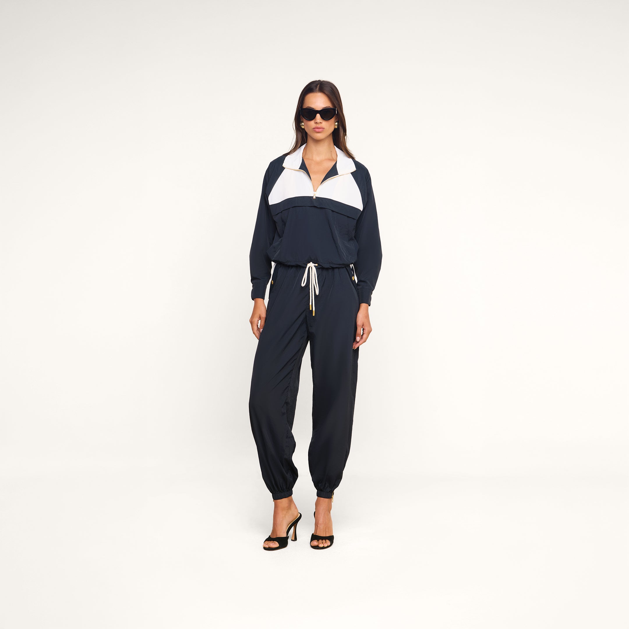 Wearing the Mariko Jogger, a navy blue and white water-repellent tracksuit with drawstring pants, a woman confidently stands against a plain white background. She accessorizes the sporty look with black heels and dark sunglasses.