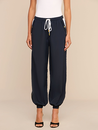 Wearing the Mariko Jogger—lightweight nylon pants with a water repellent finish and gold accents—a person pairs them with a white sleeveless top and black open-toe heels while standing against a beige background.