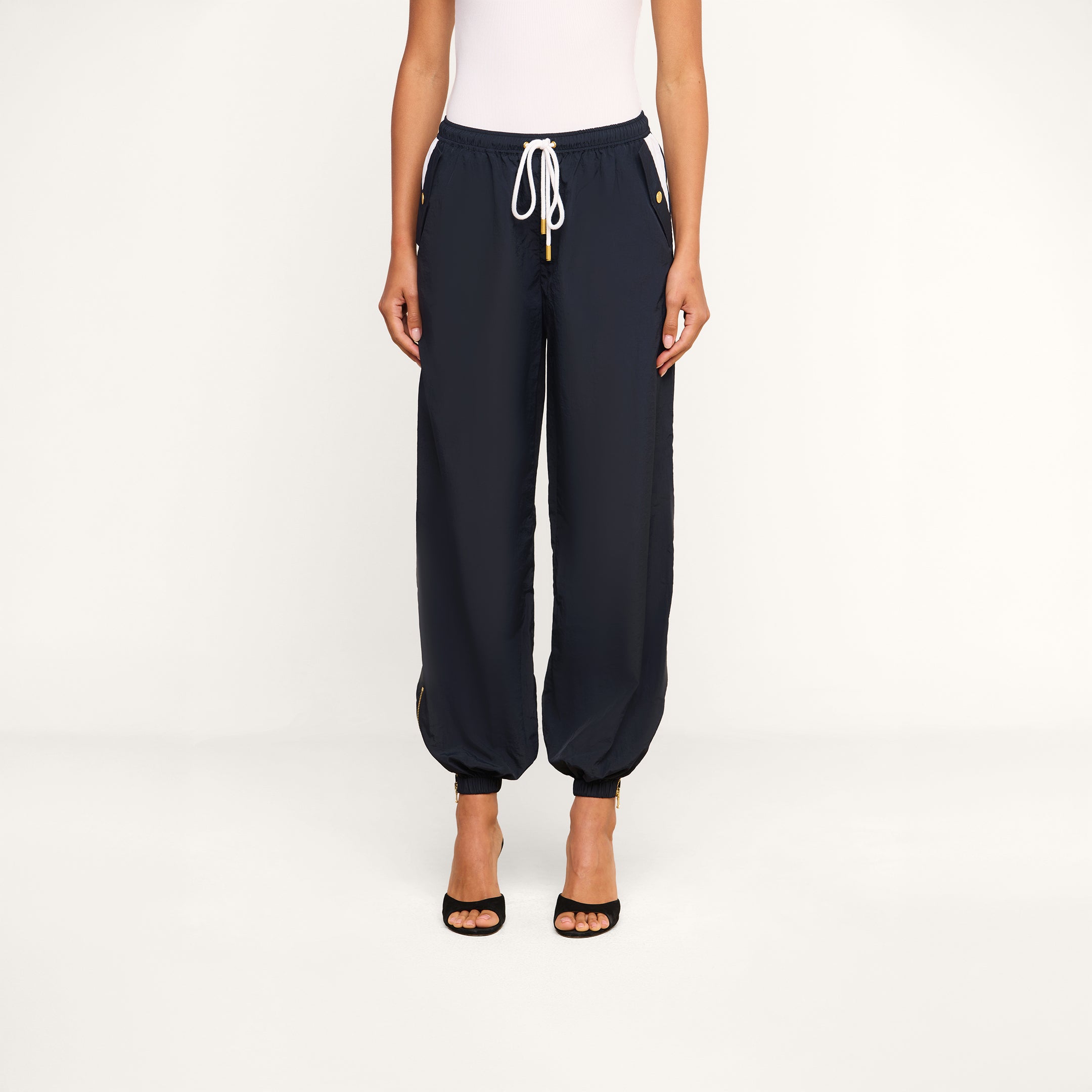 A person wearing a white sleeveless top, the Mariko Jogger—a pair of dark lightweight nylon jogger pants with zippered pockets—and black open-toe heels stands against a plain white background.