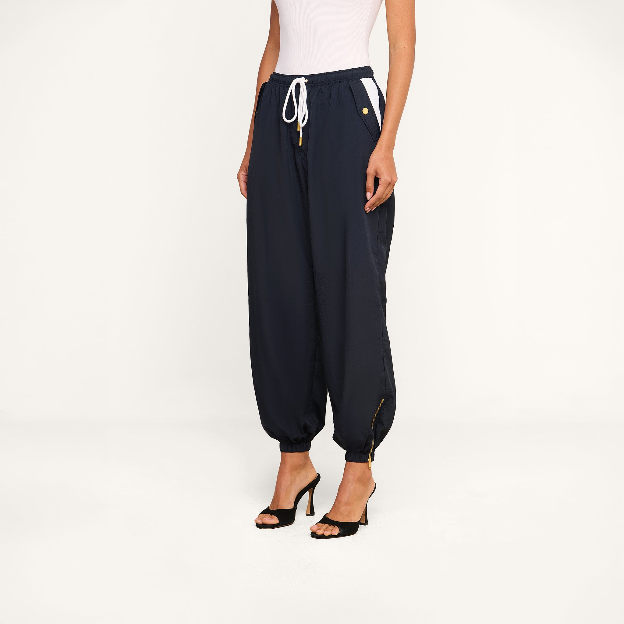 A woman wearing a white sleeveless top, the navy blue Mariko Jogger with drawstring and ankle zippers, and black high-heeled sandals stands against a plain white background.