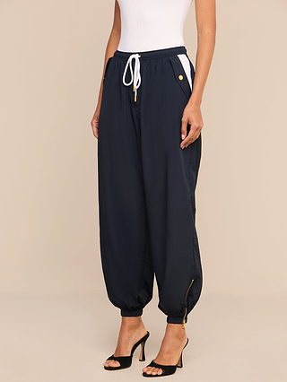 Wearing the Mariko Jogger—lightweight nylon pants with a subtle water-repellent finish, white pockets, and a drawstring—a person stands against a plain beige background, styled with a white sleeveless top and black high-heeled sandals.