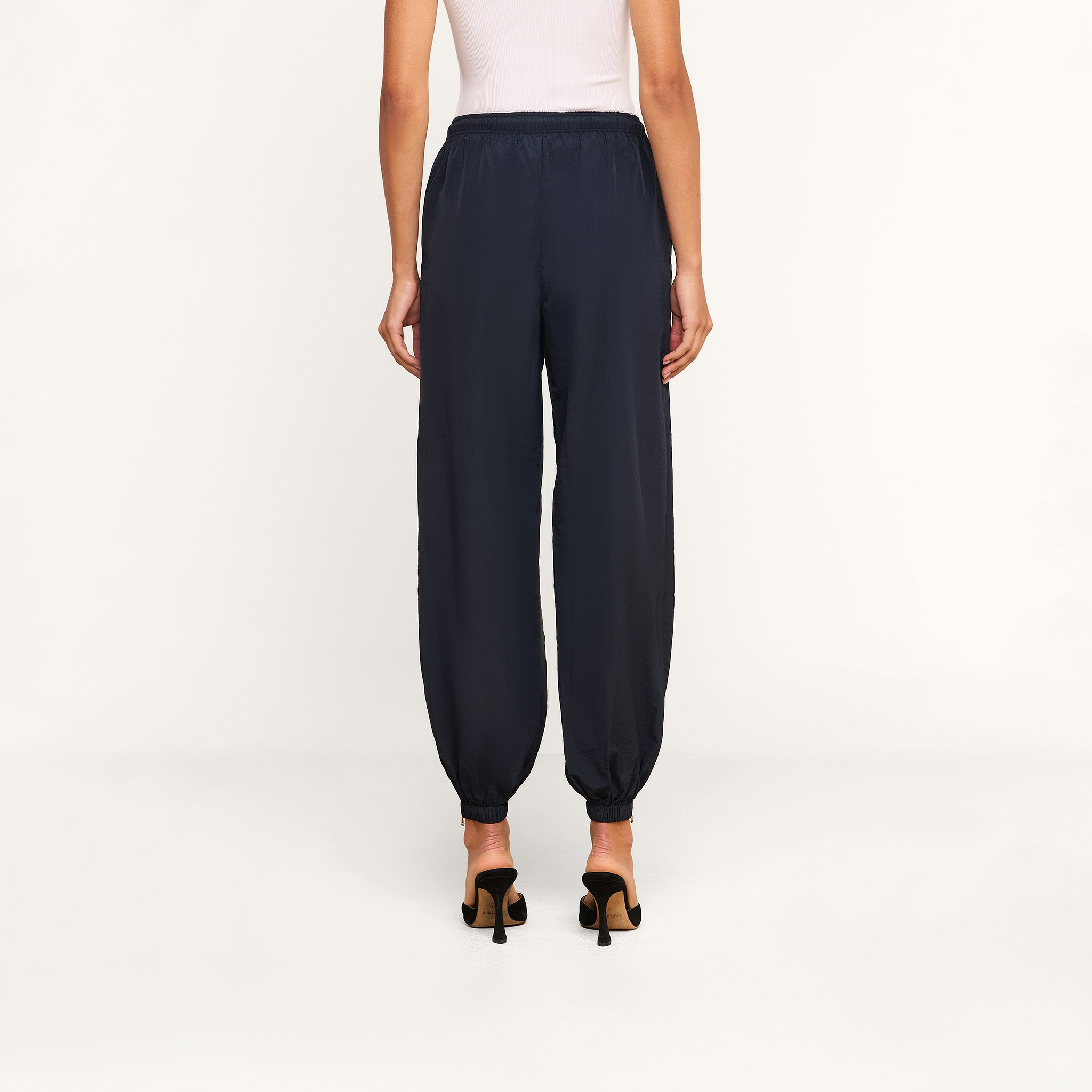 Wearing the Mariko Jogger—a pair of black, lightweight nylon jogger pants with elastic cuffs—a person stands facing away against a plain white background, styled with a white sleeveless top and black heeled sandals.