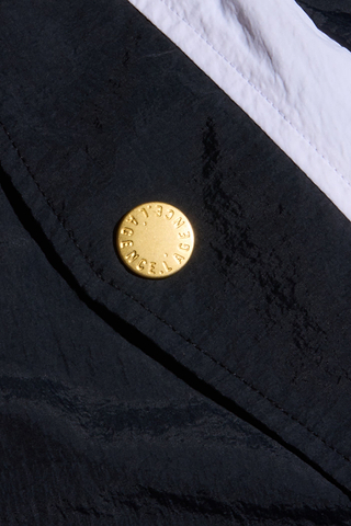 Close-up of the black lightweight nylon fabric of the Mariko Jogger, featuring a diagonal white stripe and a gold button embossed with “BALENCIAGA” in a circular pattern—a signature detail of this style.
