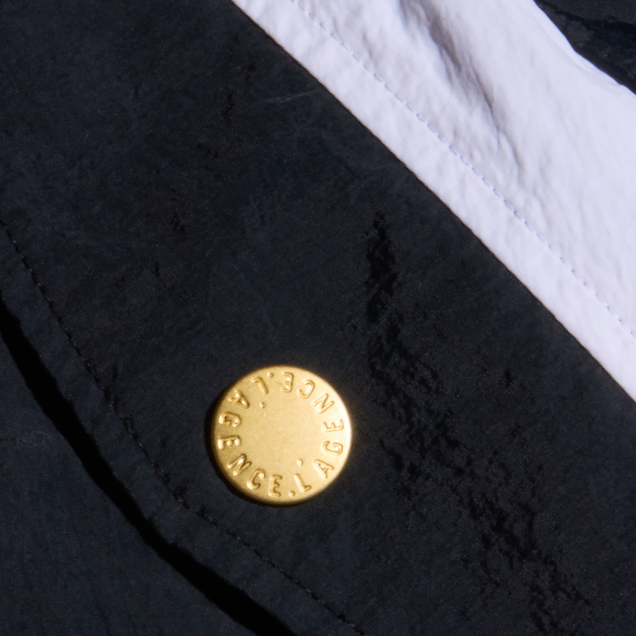 A close-up of black, water-repellent fabric from the Mariko Jogger showcases a gold button engraved with BALENCIAGA, positioned next to a white fabric seam running diagonally across the image.