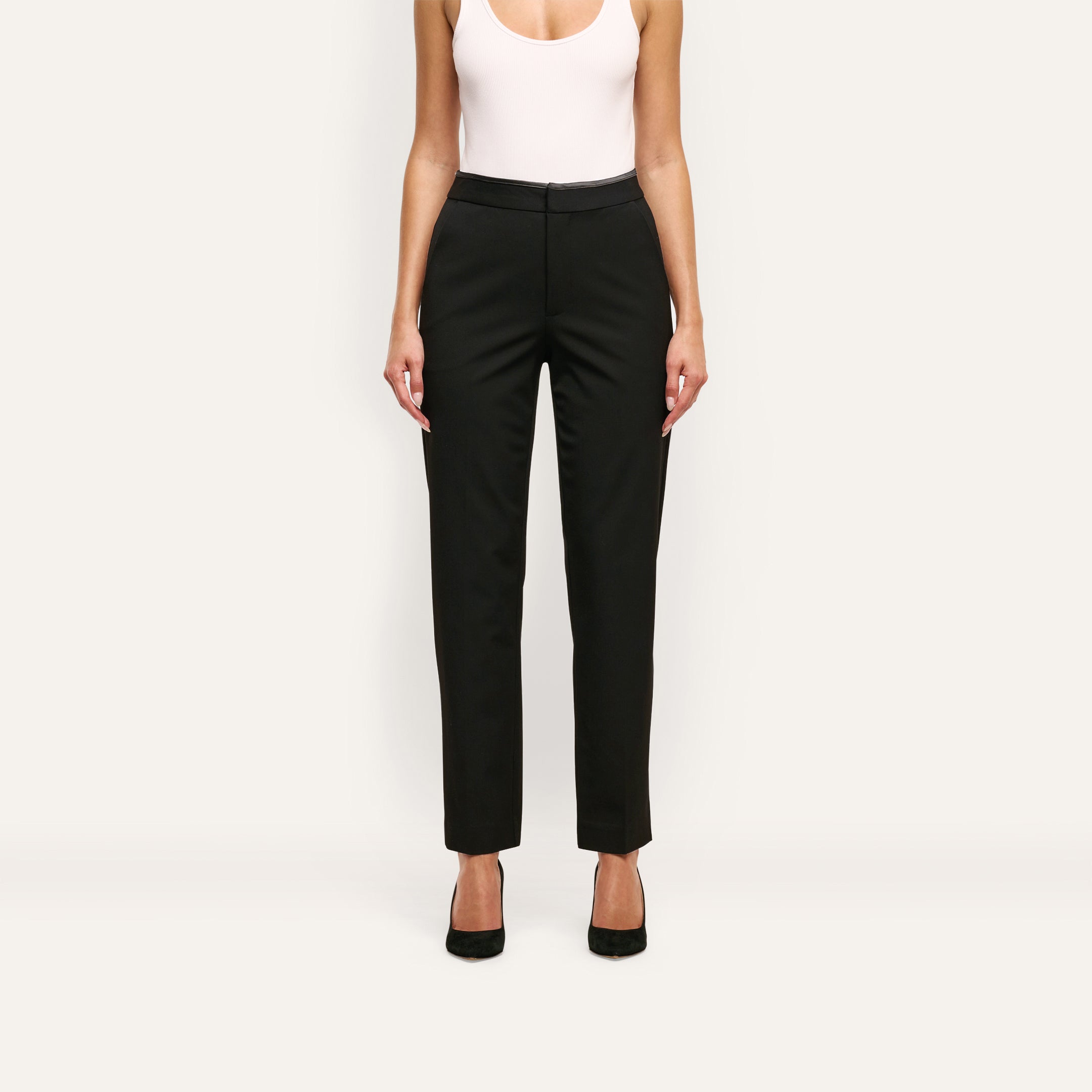 A person wearing Rebel Trouser high-waisted black stretch twill suiting trousers, a white tank top, and black pumps.
