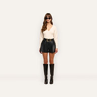 A person wearing a cream-colored V-neck sweater, Clairmont Vegan Leather Shorts with a gold-buckled belt, and black knee-high boots.