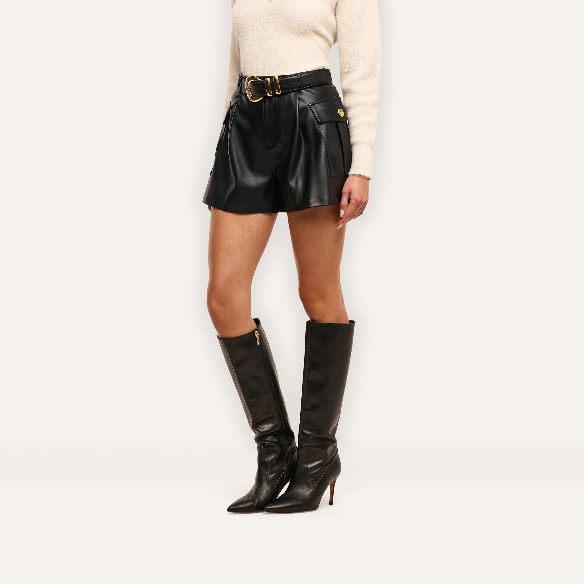 Clairmont Vegan Leather Short in black, featuring a high-waisted fit, cargo pockets, a gold-buckled belt, and paired with knee-high stiletto boots.
