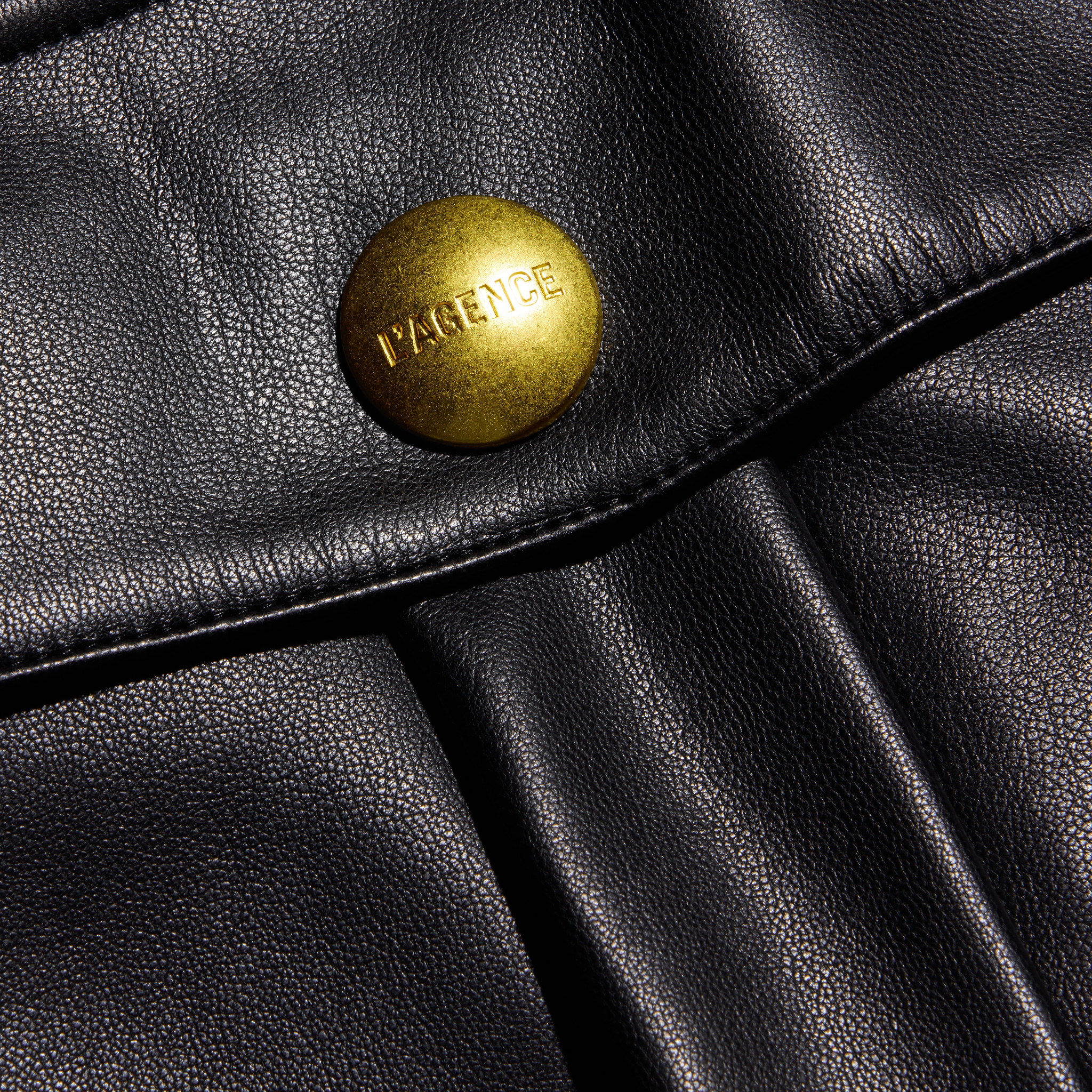 Close-up of a black vegan leather cargo pocket flap on the Clairmont Vegan Leather Short, secured by a large, round brass button embossed with the brand name.